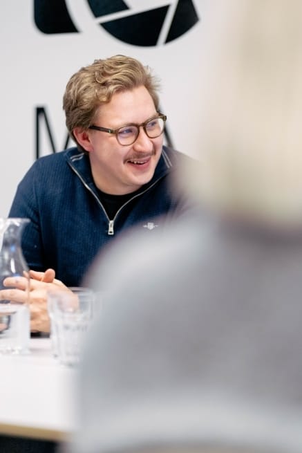 Henkilö istuu toimistossa sininen paita yllään ja nauraa, etualalla on sumennettu toinen henkilö selkä kameraan päin.