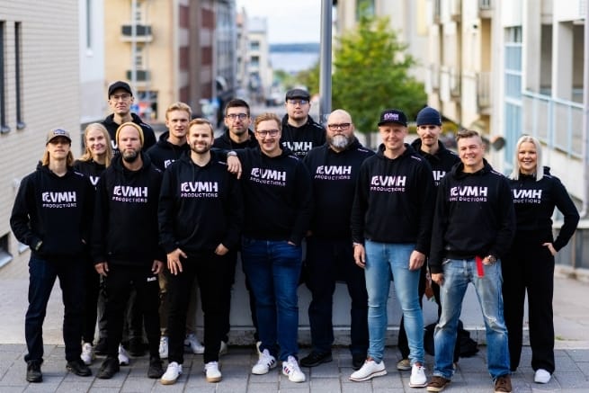 Kuvassa on useita henkilöitä, joilla on musta huppari päällään. Mustassa hupparissa on logo, jossa lukee VMH Productions. Ryhmäkuvan taustalla on puu ja rakennuksia.