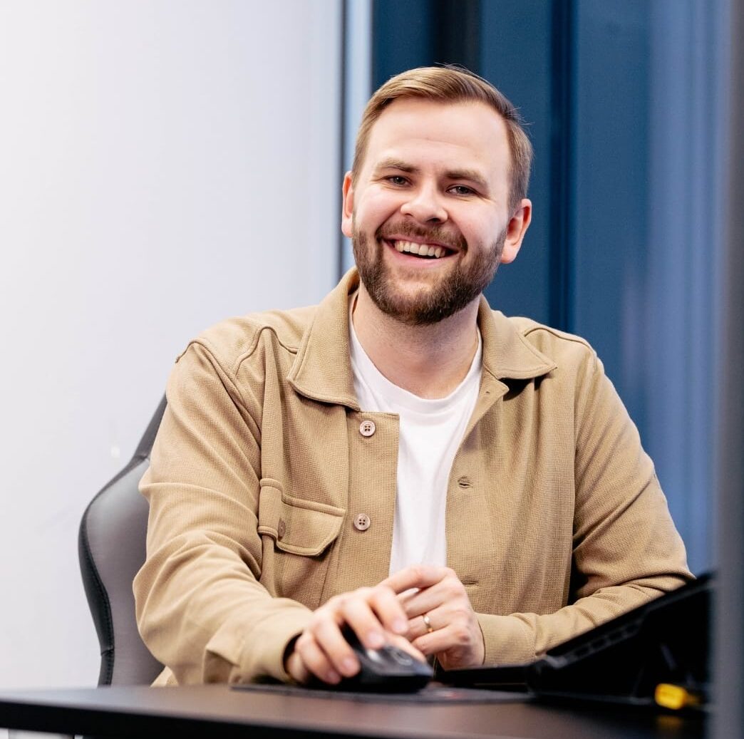 Henkilö hymyilee suoraan kameraan ja istuu työpisteellä tietokoneen hiiri kädessään yllään beige kauluspaita jonka alla on valkoinen paita. Taustallaa seinä ja sinertävät ikkunat.