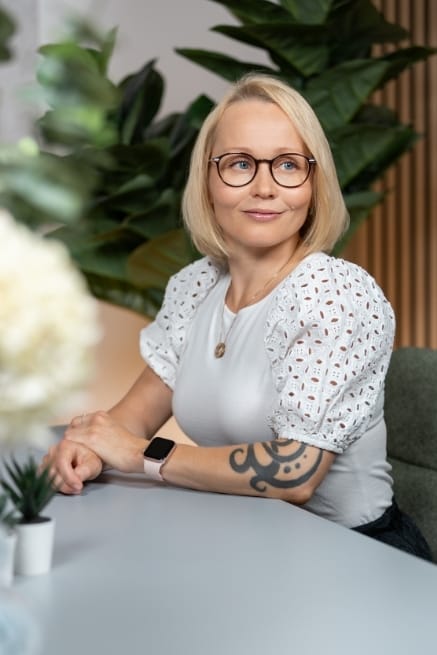 Henkilö istuu harmaan pöydän ääressä valkoisessa paidassa. Henkilöllä on kello kädessään ja päällään silmälasit. Taustalla näkyy puunväristä akustiikkalevyä sekä tekokasvi.